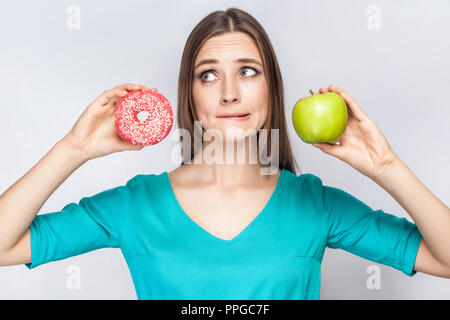 Portrait of dreaming young beautiful girl in blue blouse standing, showing and holding pink donut and green apple in hands with thoughful cunning face Stock Photo