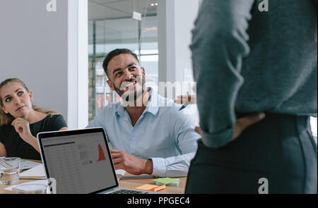 Smiling young man paying attention to the female colleagues presentation in boardroom. Happy professional in business meeting. Stock Photo