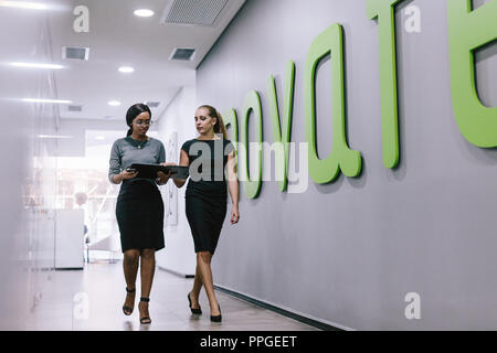Two business women walking through office corridor and looking at a file. Business professionals discussing work in modern office hallway. Stock Photo