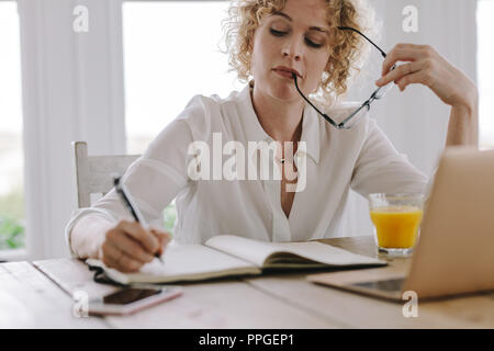 Woman working from home with laptop and juice on the table. Woman writing notes in diary holding her eyeglasses in hand. Stock Photo