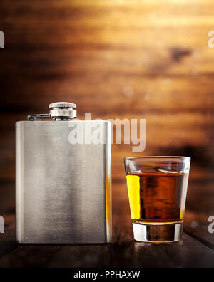 Metal whiskey hip flask closeup, on a wooden table, with a shot glass filled with whisky alcohol Stock Photo