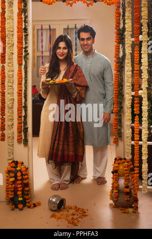 Happy young couple in traditional dress standing near the decorated entrance of their house with pooja plate. Stock Photo