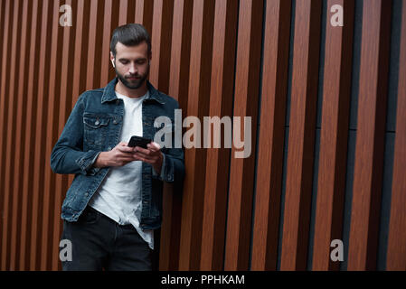 Freestyle. Young man standing leaning on wooden wall isolated browsing internet on smartphone concentrated Stock Photo