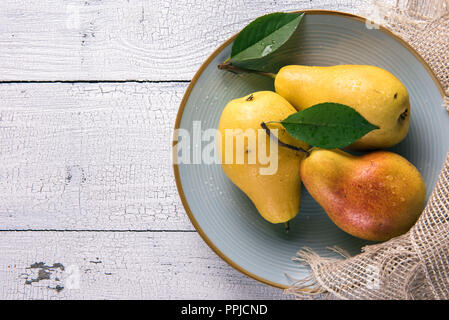 Three wet ripe yellow pears with green leaves arranged on round light blue plate on top of old white wooden table with burlap napkin from one side. Se Stock Photo