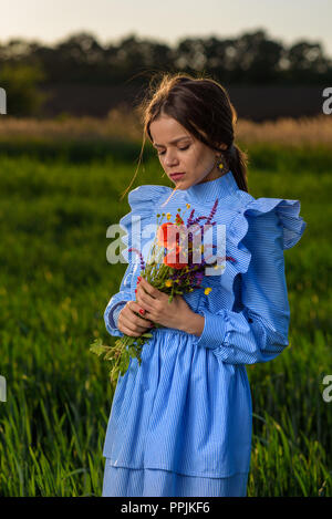Young woman in blue and white striped dress is holding with two hands a bouquet of summer flowers with her eyes closed and her head is tilted while st Stock Photo