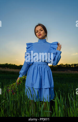 Young woman in blue and white striped dress is holding a bouquet of summer flowers with one hand while standing in the field of green wheat at a sprin Stock Photo