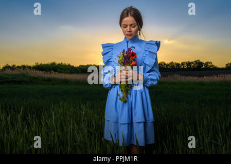 Young woman in blue and white striped dress who is holding a bouquet of summer flowers with her eyes closed and her head tilted down while standing in Stock Photo