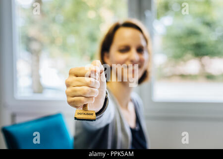 A woman in an office holds a wooden stamp with the german word 'digitalisiert' which means 'digitalized' into the camera, depth of field Stock Photo