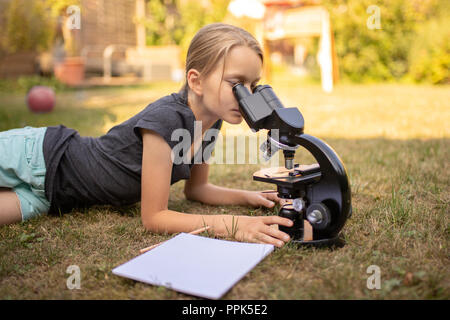 A 9 year old girl lies on the grass in the garden and looks into the eyepiece of a microscope. In front of her is a sheet of paper. Stock Photo
