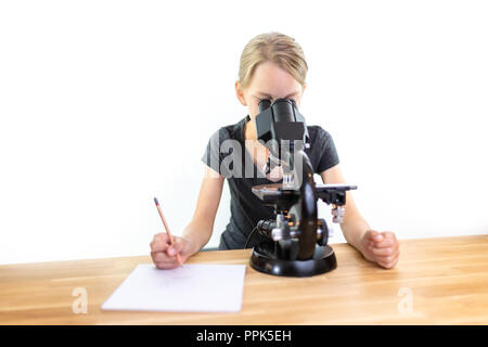 A 9-year-old girl looks into an eyepiece of a microscope and notes her observations on a sheet of paper. Isolated against white background Stock Photo