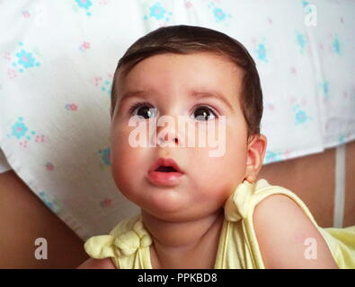 A beautiful dark haired and black eyed baby is watching in the distance, indoor at the home Stock Photo