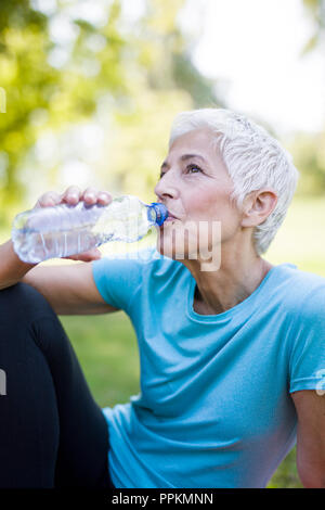 Senior woman rests and drinks water after workout in the park Stock Photo