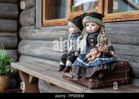Ancient porcelain dolls sits on suitcase near log farmhouse. Stock Photo