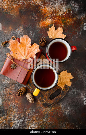 Two colored enamel tea cups, autumn maple leaves and pine cones on a rusty brown background Stock Photo