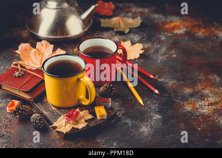 Two colored red and yellow enamel cups of tea, watercolors in cuvettes, colored pencils, autumn maple leaves and bumps on a rusty brown background Stock Photo