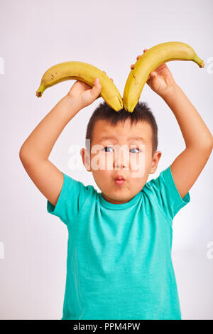 Child boy in a turquoise shirt, holds bananas depicting horns and looks with eyes flattened - fruit and healthy food Stock Photo