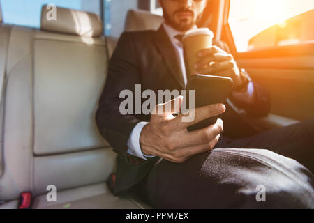 cropped image of businessman sitting in car with disposable coffee cup and using smartphone Stock Photo