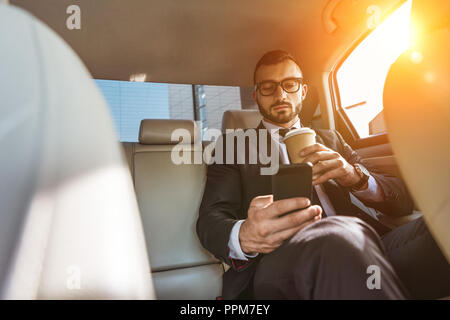 handsome businessman sitting in car with disposable coffee cup and using smartphone Stock Photo