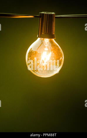 An outdoor lightbulb, glowing yellow, hanging on a black cord in early evening Stock Photo