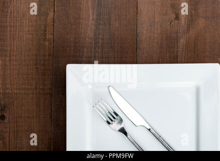 Square white dinner plate on a wooden table with fork and knife. Stock Photo