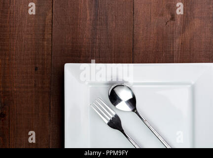 Square white dinner plate on a wooden table with fork and spoon. Stock Photo