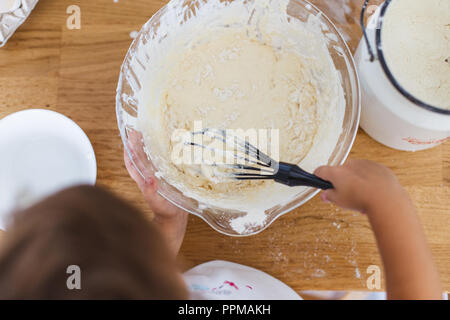 Little girl preparing dough for pancakes at the kitchen. Concept of food preparation, white kitchen on background. Casual lifestyle photo series in re