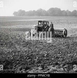 1950s, historical, in the middle of a rough ground field, a farmer stands on the back of his tractor, attached with a soil raking machine,  England, UK. Stock Photo