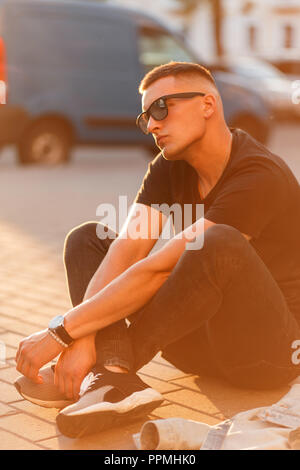 Stylish hipster man with sunglasses in black fashion clothes is sitting on the street at sunset Stock Photo