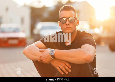 Stylish handsome young man with sunglasses in trendy black clothes sits asphalt at sunset Stock Photo