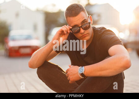 Fashionable handsome young man with sunglasses in a black T-shirt is sitting on the street at sunset Stock Photo