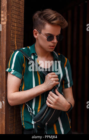 Fashionable handsome model with a sunglasses in a beach fashion shirt and bag stands near a wooden wall Stock Photo