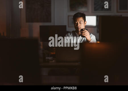 Young Asian businessman working at his desk in an office late at night drinking a cup of coffee to stay awake Stock Photo