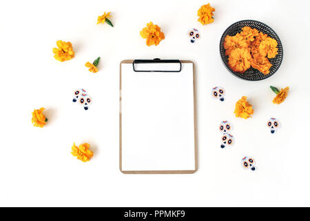 Dia de los Muertos, Mexican Day of the Dead composition. Clipboard, blank paper mockup, orange tagetes, marigold flowers and decorative paper skull stickers. White table background. Flat lay, top view Stock Photo