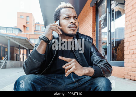 Dark-skinned pensive man talking by mobile phone and sitting on steps. Stock Photo