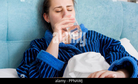 Portrait of sick young woman lying in bed and drinking water from glass Stock Photo