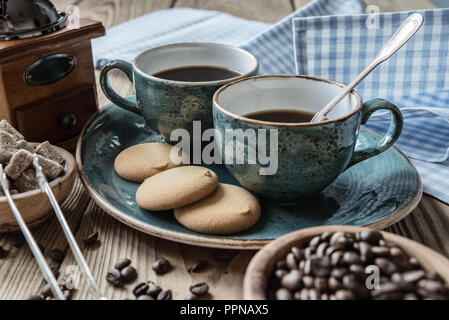 Two blue cups of black coffee, cookies and sugar pieces surrounded by checkered linen cloth and coffee beans on old wooden table Stock Photo