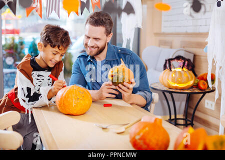 Handsome dark-haired boy wearing skeleton suit coloring pumpkin with father Stock Photo