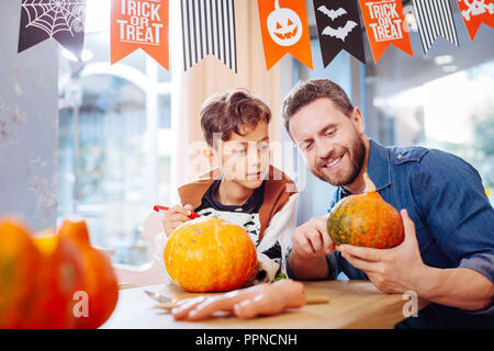 Dark-haired boy wearing skeleton costume watching his father carving pumpkin Stock Photo