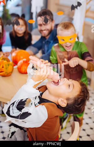 Dark-haired funny boy wearing skeleton Halloween costume eating hand cookie Stock Photo