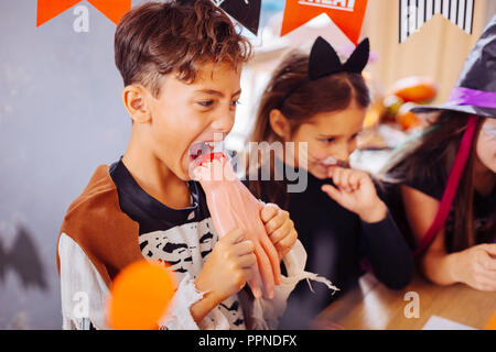 Dark-haired funny boy wearing Halloween costume eating hand cookie Stock Photo
