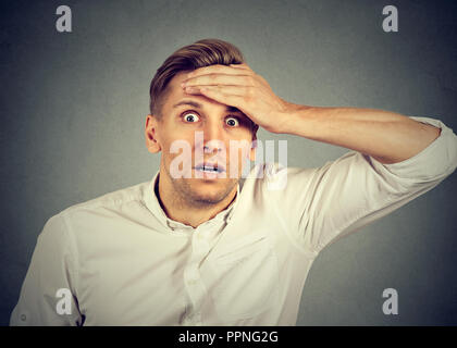 Adult man in white shirt holding hand on forehead and looking super amazed at camera on gray background Stock Photo