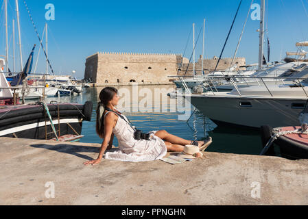 Happy tourist girl on holiday trip to Heraklion, Crete, Greece. Young woman traveler enjoys sunshine sitting on the dock of Heraklion Venetian port marina. Stock Photo