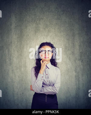 Young brunette woman in eyeglasses looking up in contemplation on gray background Stock Photo