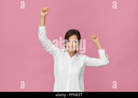 Smiling positive female with victory gesture and attractive look, wearing white shirt, posing against pink background Stock Photo