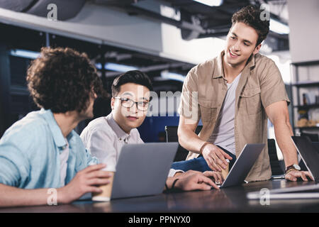 three multicultural businessmen having meeting with paper cups of coffee at table with laptops in modern office Stock Photo