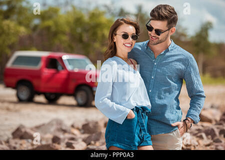 young smiling couple in sunglasses hugging together, car on background Stock Photo