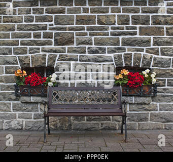 Wooden bench in front of old gray stone wall with two small windows decorated with colorful flowers Stock Photo