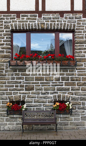 Wooden bench in front of old gray stone wall with two small windows decorated with colorful flowers Stock Photo