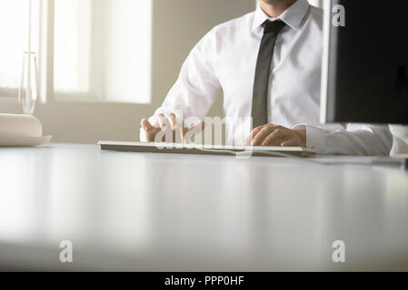 Businessman sitting typing on a wired computer keyboard in a low angle view of his hands on the desk past the monitor and copy space. Stock Photo