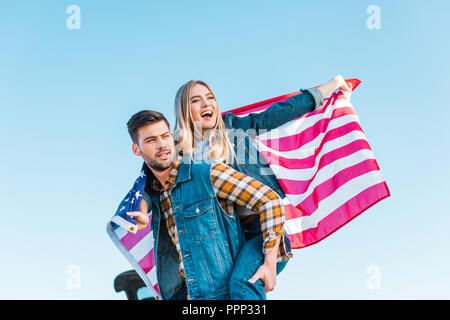 stylish man doing piggyback ride to girlfriend holding american flag against blue sky, independence day concept Stock Photo